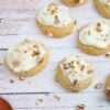 Dairy-free collagen carrot cookies topped with cream cheese frosting and pecans on a piece of parchment paper with shredded carrots beside them.