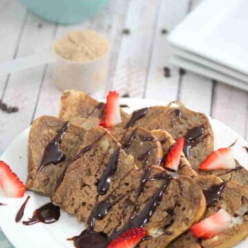 Chocolate protein French toast on a white plate drizzled with chocolate and strawberries with the plate on a blue linen.