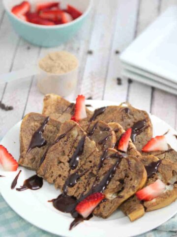 Chocolate protein French toast on a white plate drizzled with chocolate and strawberries with the plate on a blue linen.