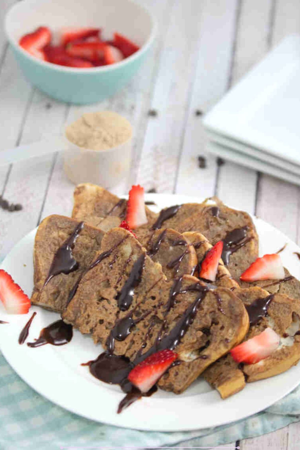 Chocolate protein French toast on a white plate drizzled with chocolate and strawberries with the plate on a blue linen.
