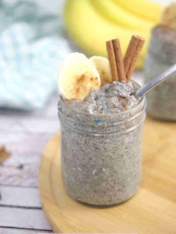 Banana chia pudding in a mason jar with a spoon in it and sliced bananas, and cinnamon on top and the jar on a wooden board.