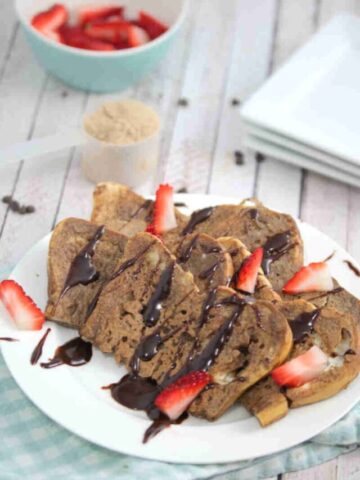 Chocolate protein French toast on a white plate drizzled with chocolate and strawberries with the plate on a blue linen.