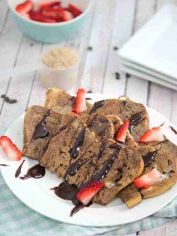 Chocolate protein French toast on a white plate drizzled with chocolate and strawberries with the plate on a blue linen.