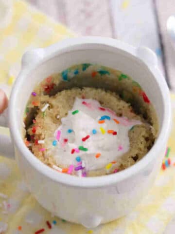 Hand holding a white mug on a yellow linen with a protein mug cake in it topped with dairy-free whip cream and sprinkles.