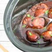 Slow cooker BBQ chicken thighs in a black slow cooker with green onions on top and one thigh on a spoon being pulled out.