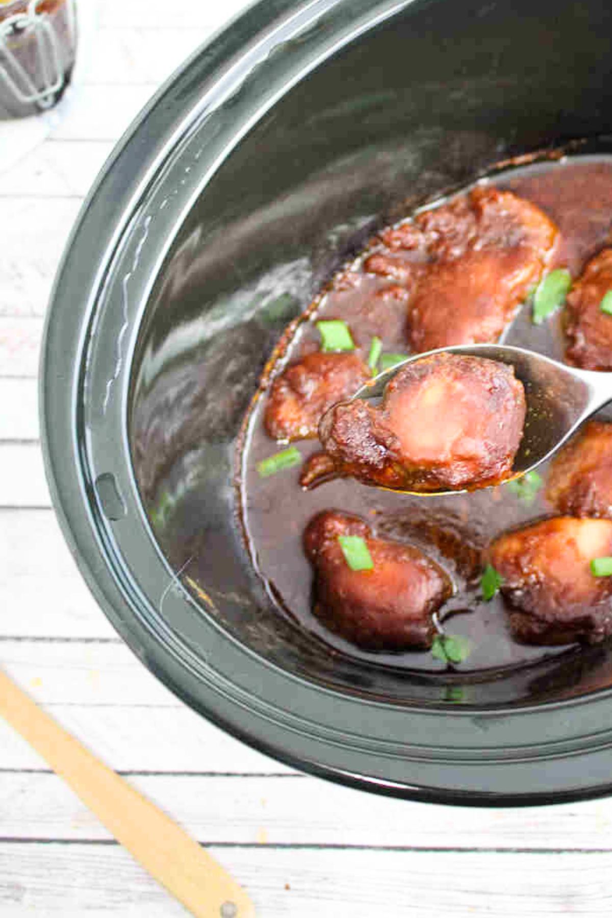 Slow cooker BBQ chicken thighs in a black slow cooker with green onions on top and one thigh on a spoon being pulled out.