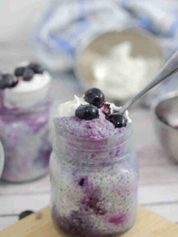 Blueberry chia pudding in a small mason jar on a cutting board with a spoon in the jar.