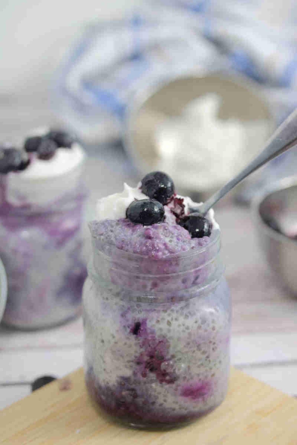 Blueberry chia pudding in a small mason jar on a cutting board with a spoon in the jar.