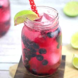 Blueberry mocktail in a mason jar with limes on the side of the jar and red straws with the jar on a wooden coater and cutting board and ingredients around it.