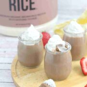 Three glass jars of chocolate protein mousse topped with coconut whipped cream on a wooden cutting board with strawberries around them and a spoonful in front.