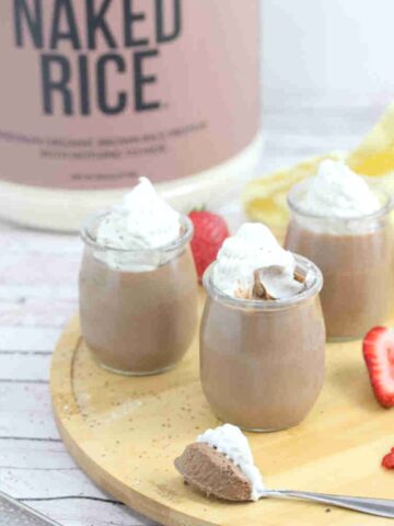 Three glass jars of chocolate protein mousse topped with coconut whipped cream on a wooden cutting board with strawberries around them and a spoonful in front.