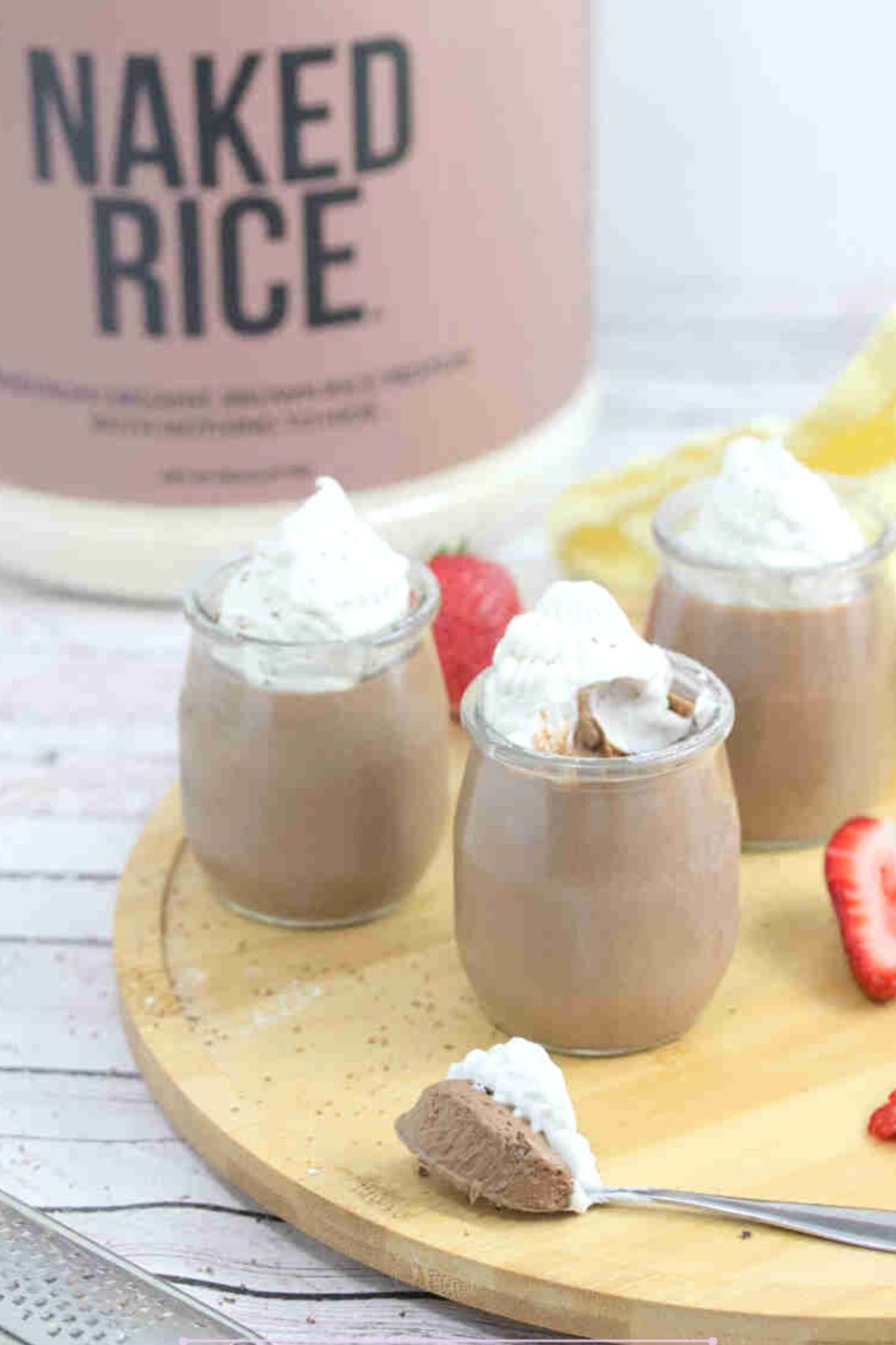 Three glass jars of chocolate protein mousse topped with coconut whipped cream on a wooden cutting board with strawberries around them and a spoonful in front.