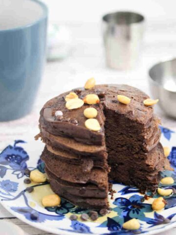Blue and white plate with chocolate protein pancakes topped with peanuts with a coffee mug and silver bowl behind them.