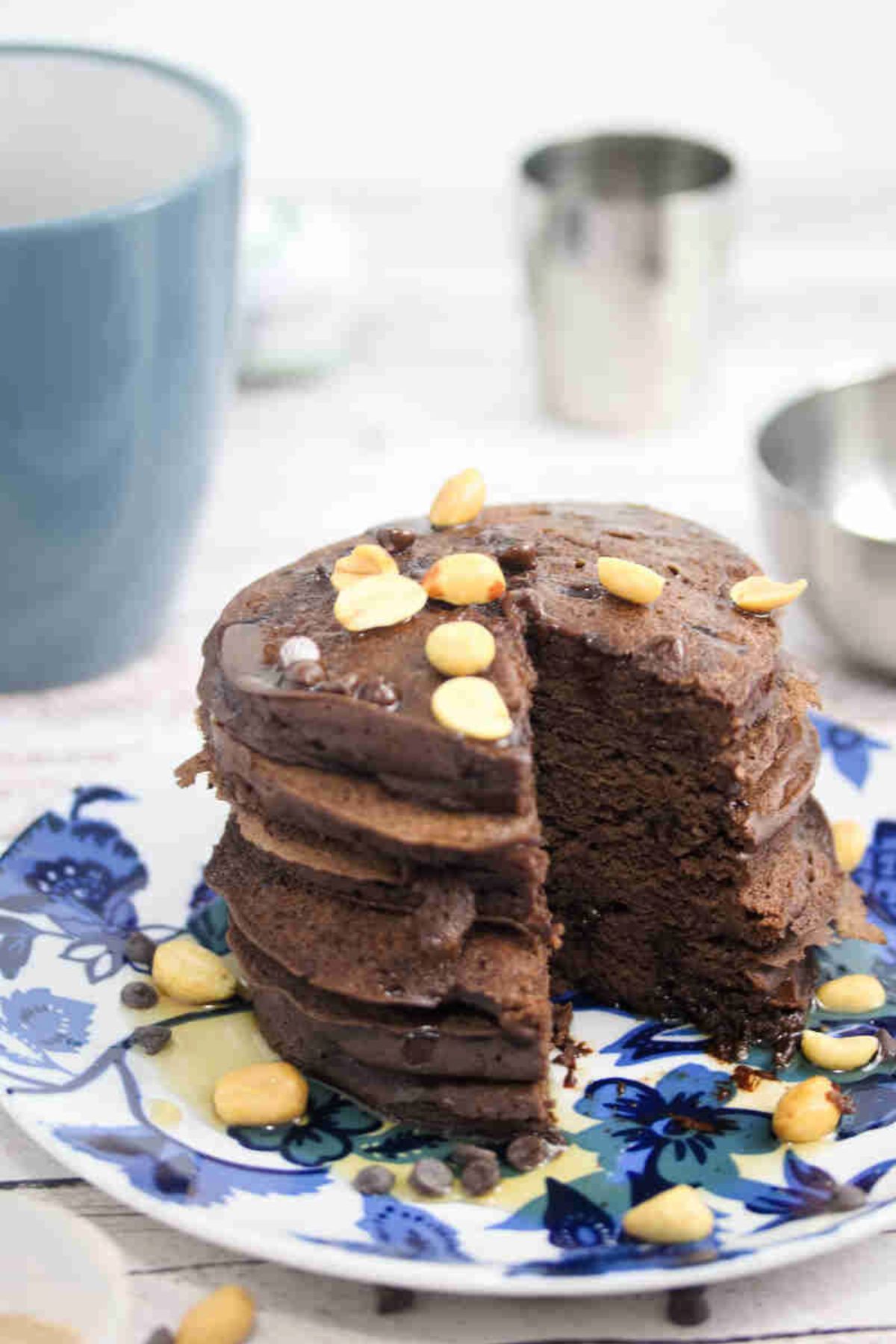 Blue and white plate with chocolate protein pancakes topped with peanuts with a coffee mug and silver bowl behind them.