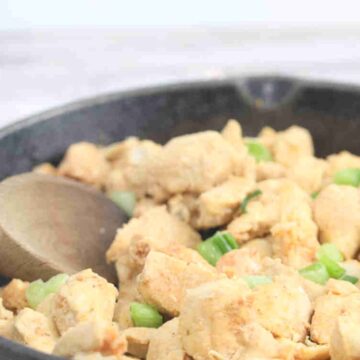 Spicy chicken bites in a cast iron skillet with green onions on top and a wooden spoon in the pan.