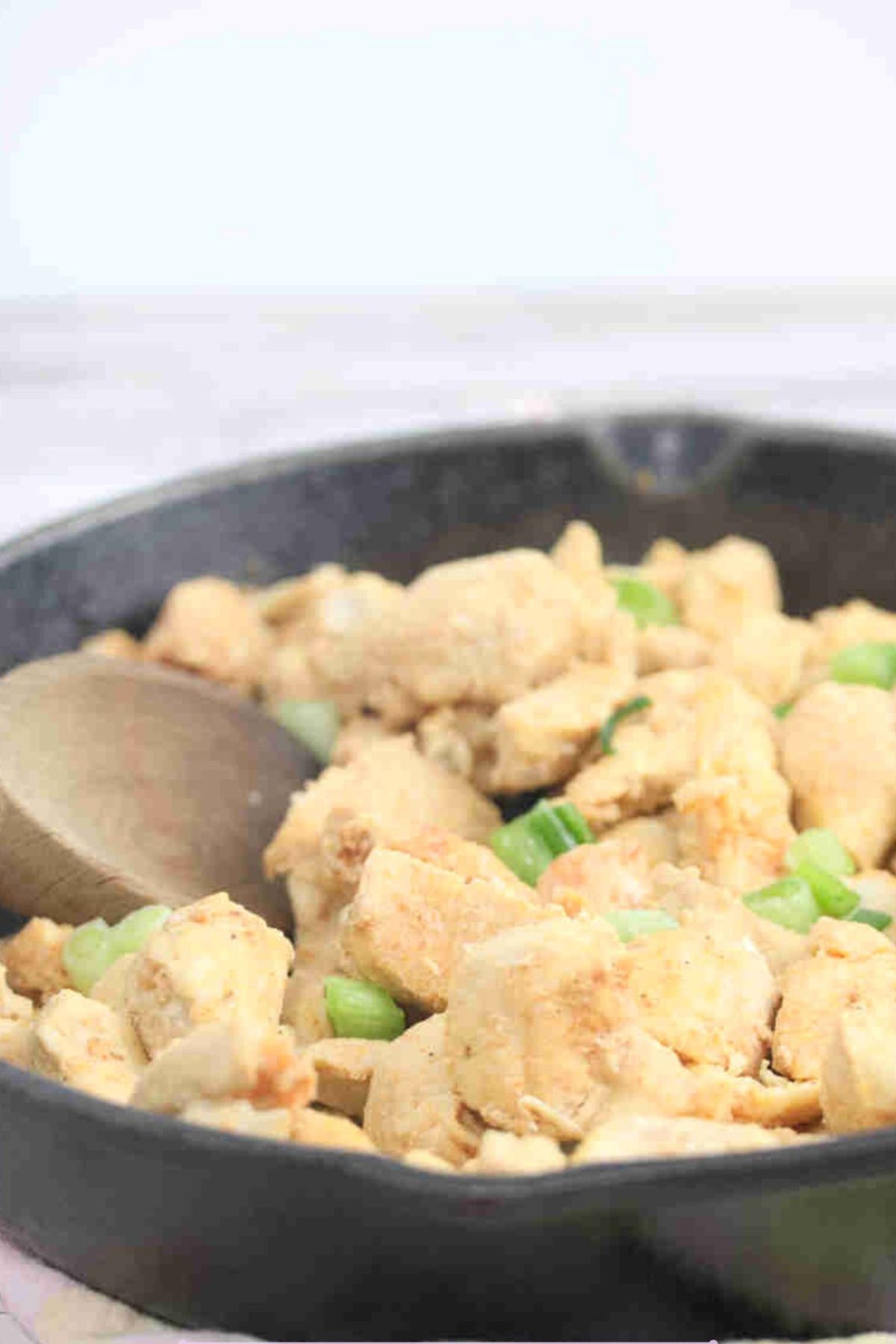 Spicy chicken bites in a cast iron skillet with green onions on top and a wooden spoon in the pan.