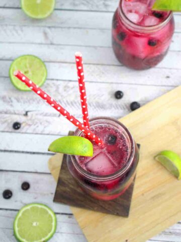 Blueberry mocktail in a mason jar with limes on the side of the jar and red straws with the jar on a wooden coater and cutting board and ingredients around it.