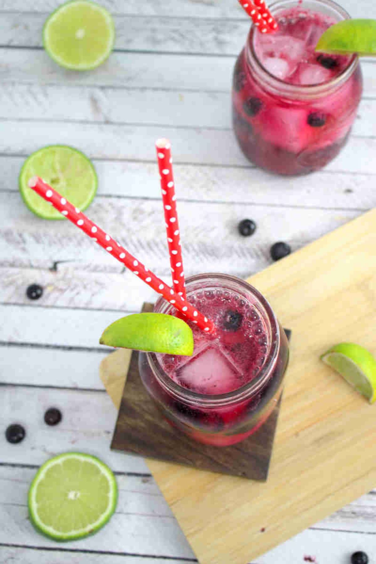 Blueberry mocktail in a mason jar with limes on the side of the jar and red straws with the jar on a wooden coater and cutting board and ingredients around it.