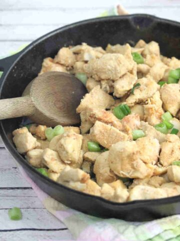 Spicy chicken bites in a cast iron skillet with green onions on top and a wooden spoon in the pan.