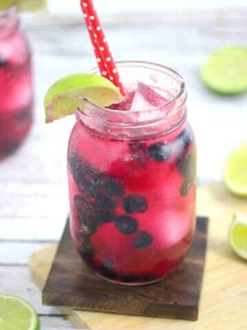 Blueberry mocktail in a mason jar with limes on the side of the jar and red straws with the jar on a wooden coater and cutting board and ingredients around it.