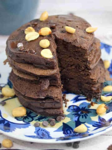 Blue and white plate with chocolate protein pancakes topped with peanuts with a coffee mug and silver bowl behind them.