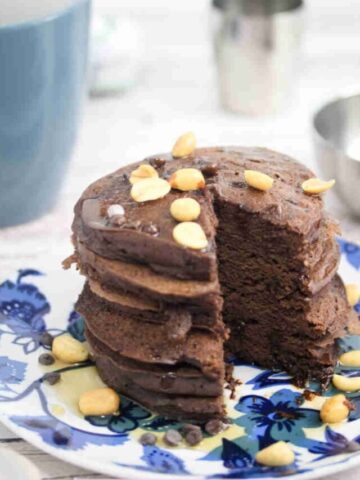 Blue and white plate with chocolate protein pancakes topped with peanuts with a coffee mug and silver bowl behind them.