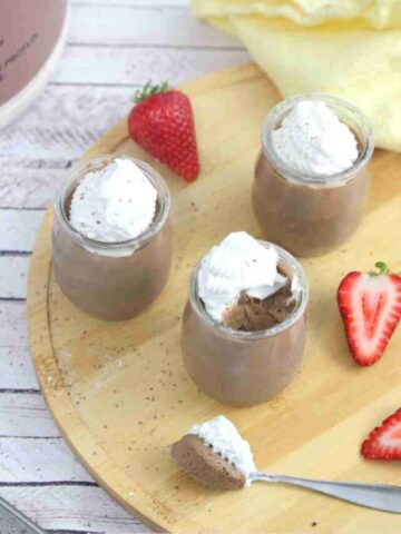 Three glass jars of chocolate protein mousse topped with coconut whipped cream on a wooden cutting board with strawberries around them and a spoonful in front.