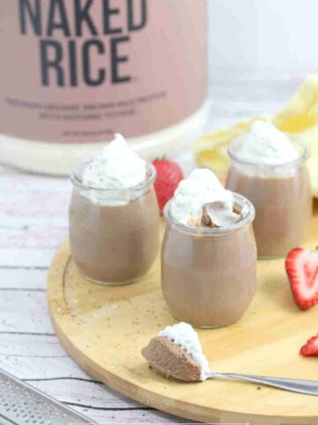 Three glass jars of chocolate protein mousse topped with coconut whipped cream on a wooden cutting board with strawberries around them and a spoonful in front.