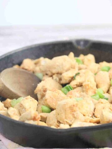 Spicy chicken bites in a cast iron skillet with green onions on top and a wooden spoon in the pan.