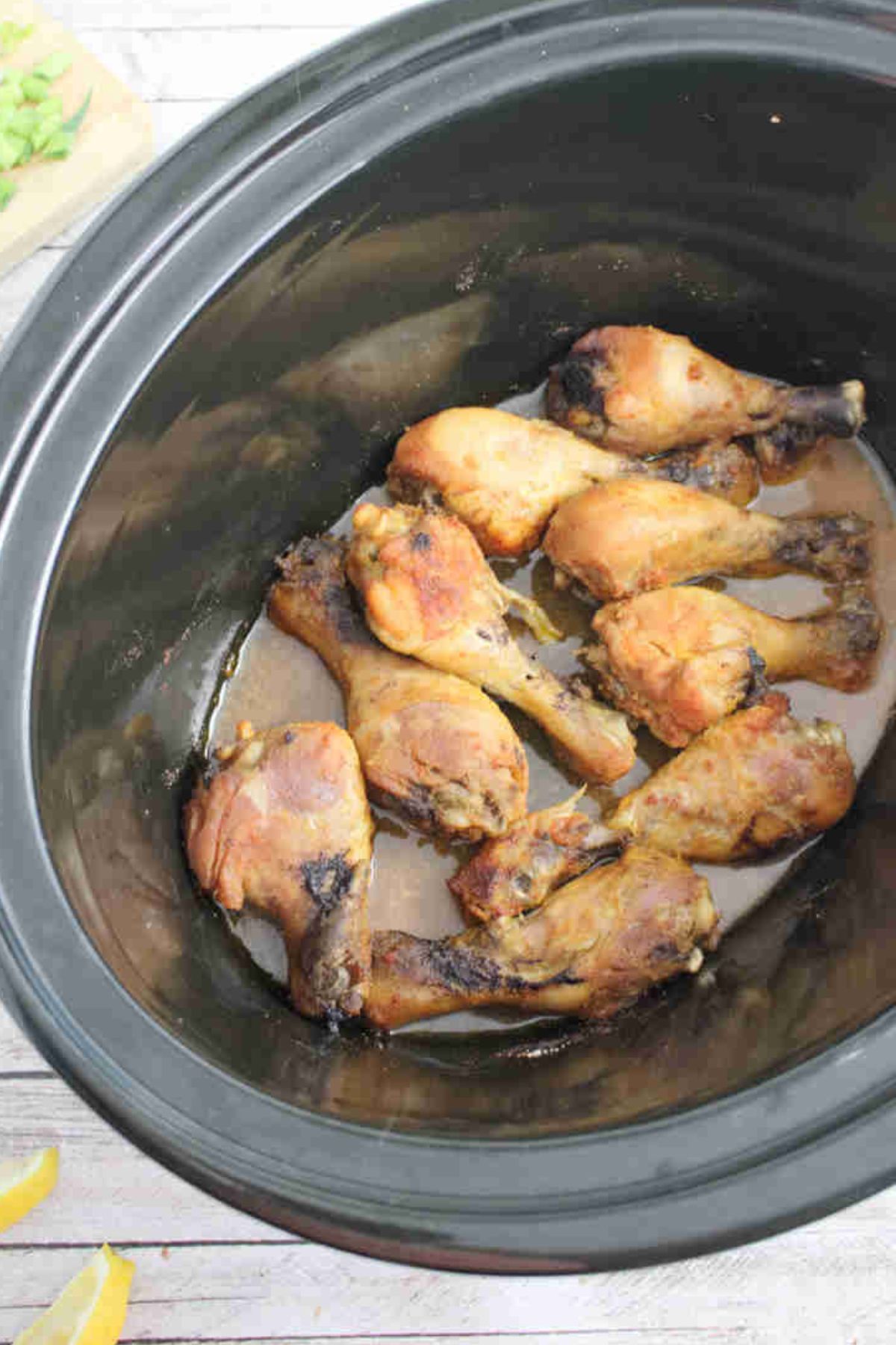 Dry rub drumsticks in a black slow cooker with a cutting board and green onions behind them.