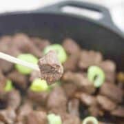 Close up of a steak bite on a wooden spear with more in a pan behind it.