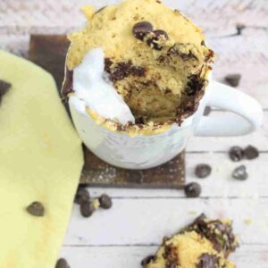 Chocolate chip mug cake with coconut cream in a white mug on a dark coasted with a yellow linen by it.
