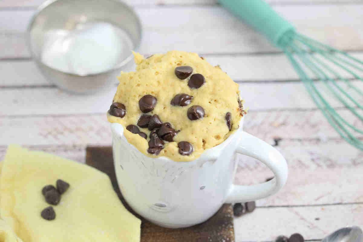 white mug with chocolate chip mug cake in it on a wooden cutting board and a whisk and coconut cream behind it.