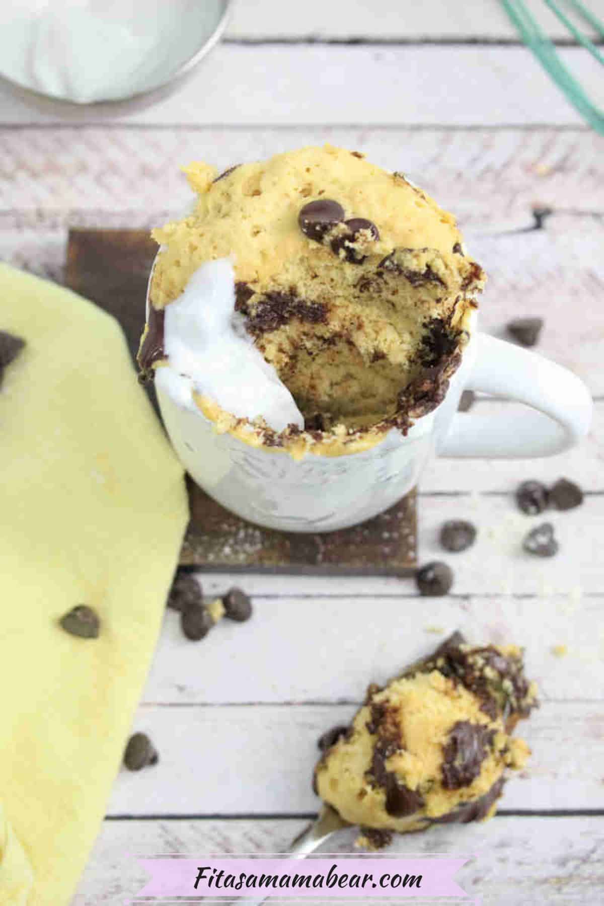 Chocolate chip mug cake with coconut cream in a white mug on a dark coasted with a yellow linen by it.