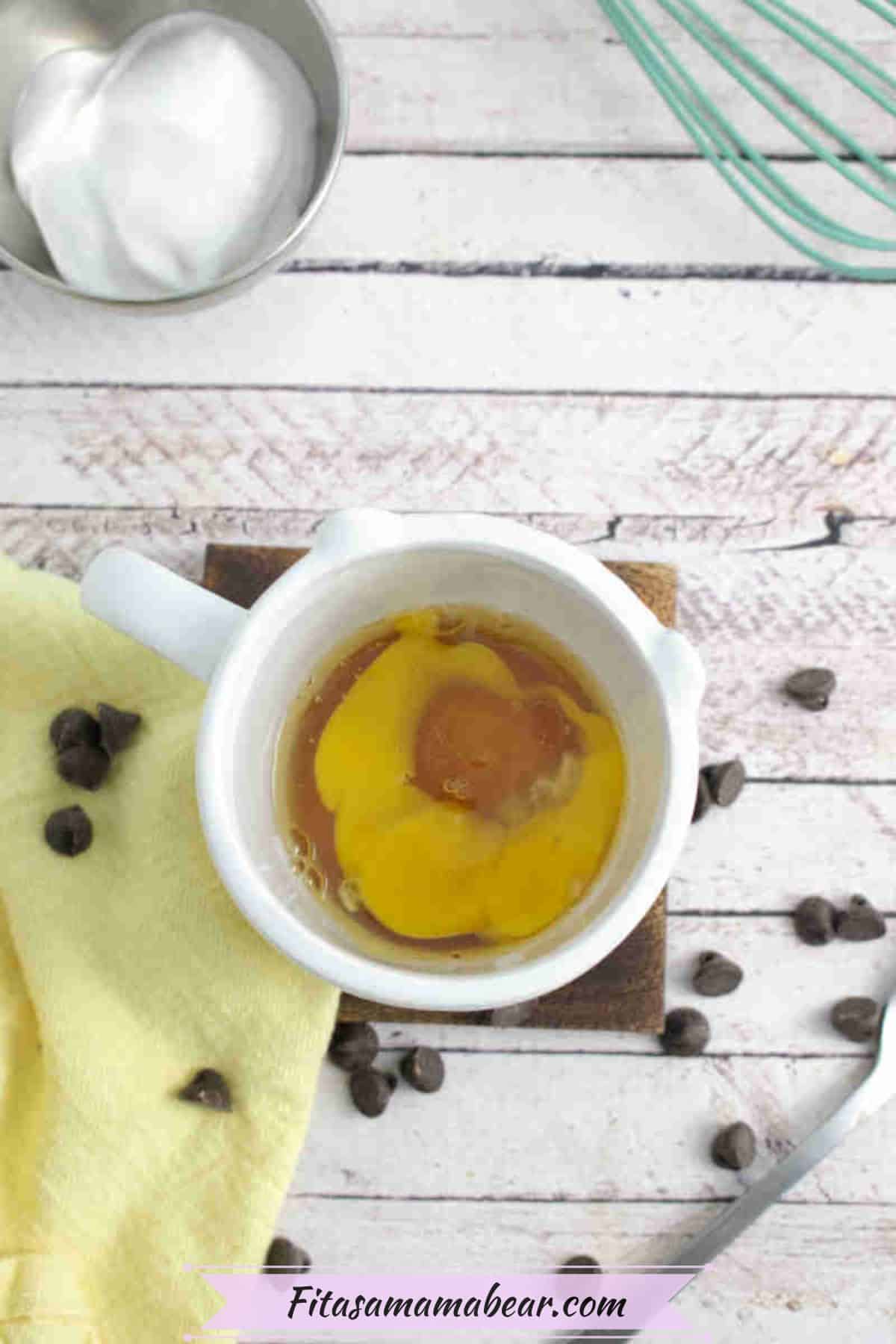 White mug with raw eggs and maple syrup in it with chocolate chips and a yellow linen beside the mug.