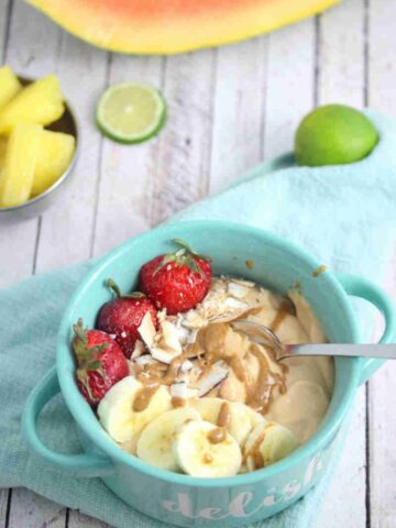 Papaya smoothie bowl topped with berries, banana and coconut in a blue bowl with a spoon and fresh fruit behind it.