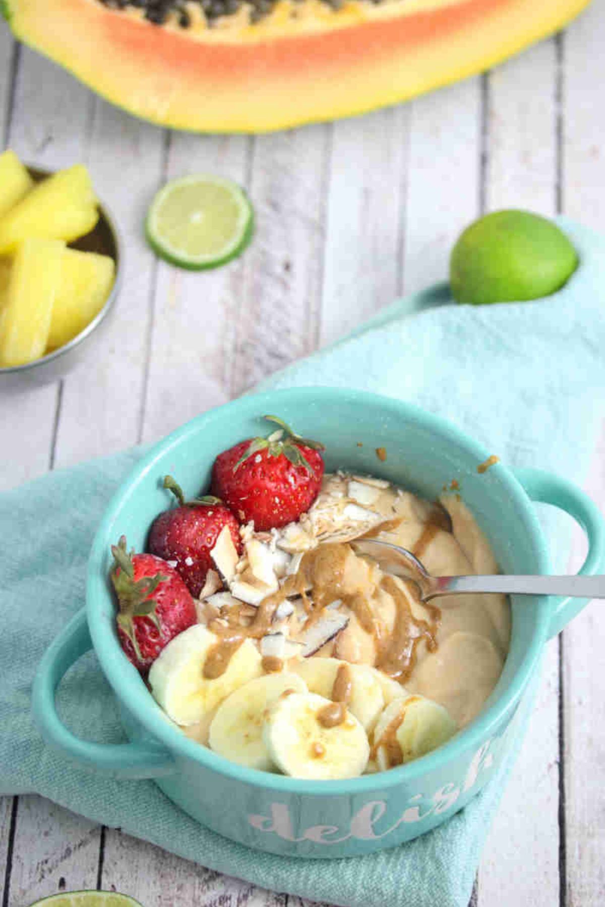 Papaya smoothie bowl topped with berries, banana and coconut in a blue bowl with a spoon and fresh fruit behind it.