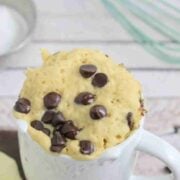 White mug with chocolate chip mug cake in it on a wooden cutting board and a whisk and coconut cream behind it.