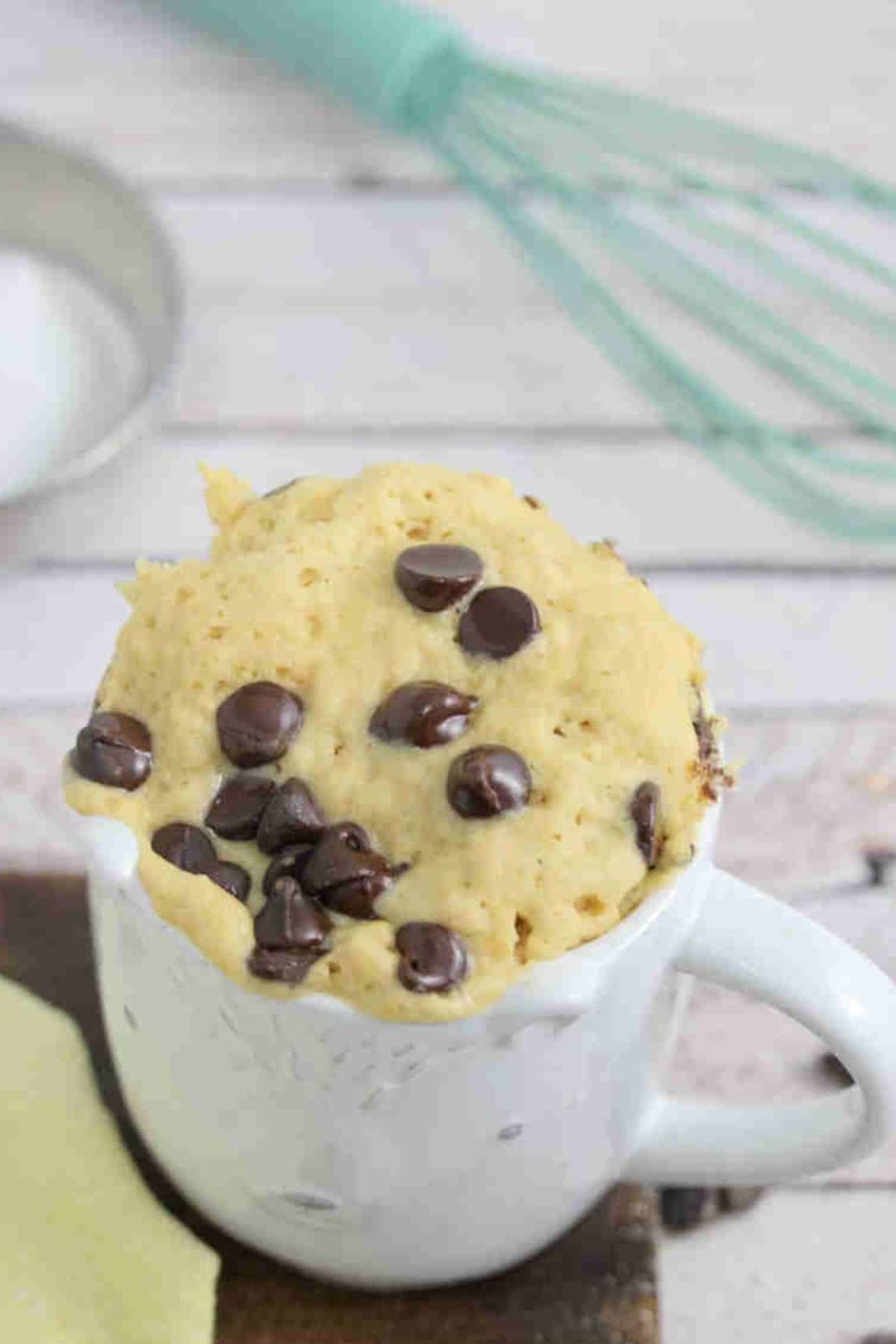 White mug with chocolate chip mug cake in it on a wooden cutting board and a whisk and coconut cream behind it.