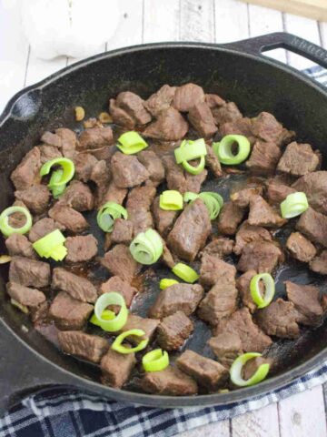 Teriyaki steak bites in a cast iron pan with leeks on top on a blue linen.