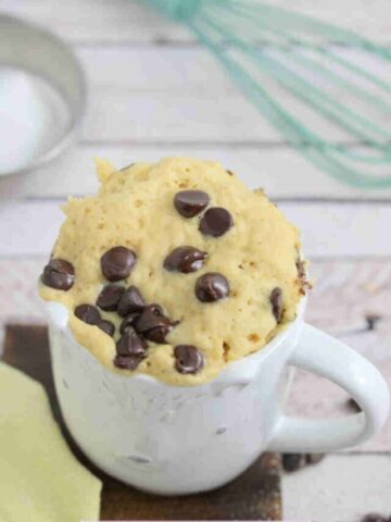 white mug with chocolate chip mug cake in it on a wooden cutting board and a whisk and coconut cream behind it.