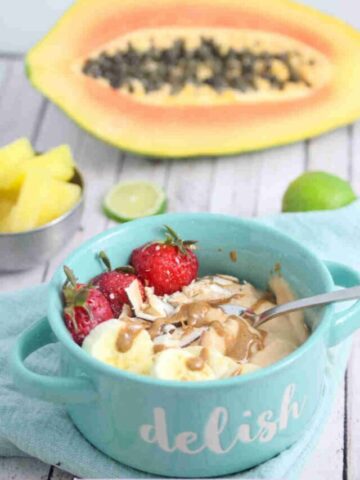 Papaya smoothie bowl topped with berries, banana and coconut in a blue bowl with a spoon and fresh fruit behind it.