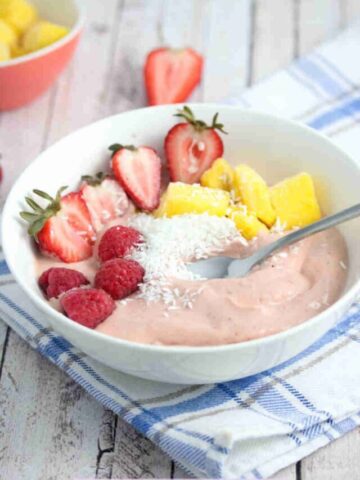 Strawberry smoothie bowl topped with berries, mango, and coconut in a white bowl on a blue linen with fruit behind it.