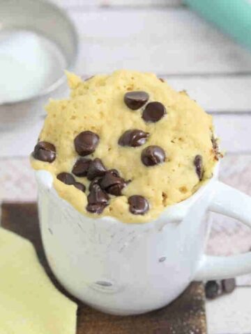 white mug with chocolate chip mug cake in it on a wooden cutting board and a whisk and coconut cream behind it.