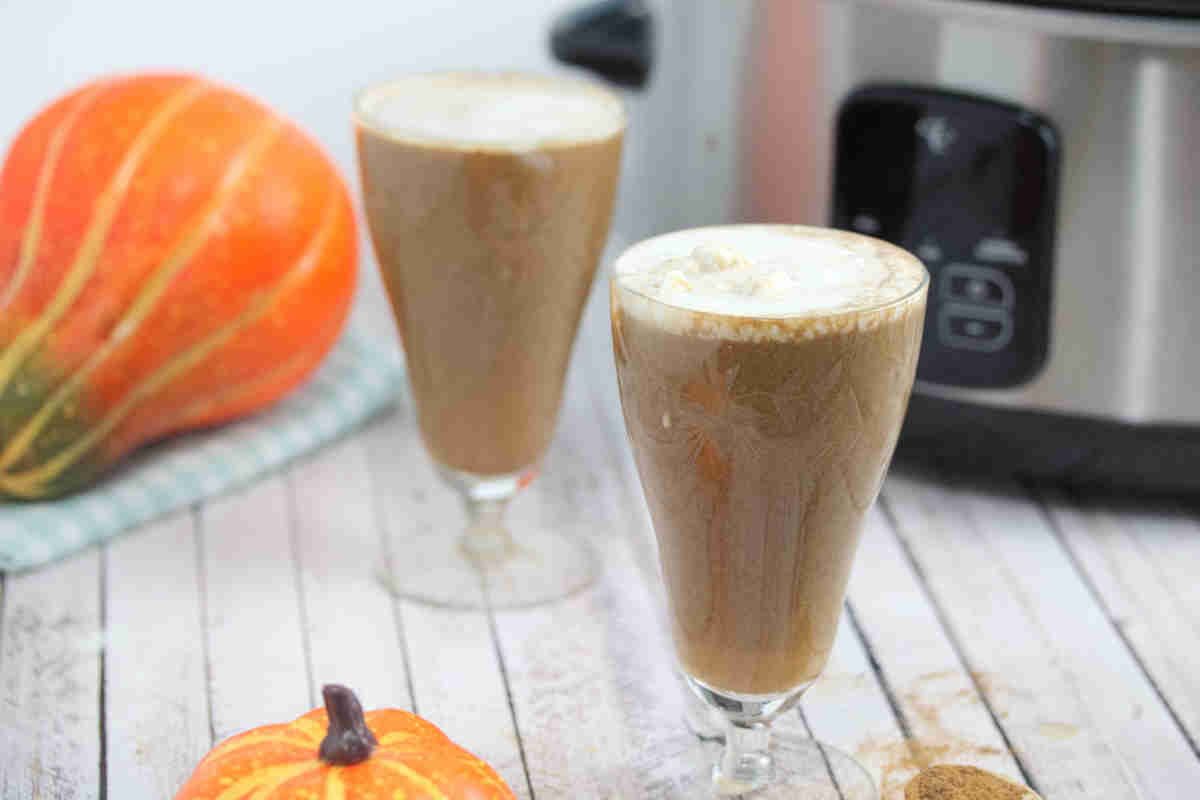 Two tall glasses with slow cooker pumpkin spice latte topped with coconut milk and pumpkins and a slow cooker around them.