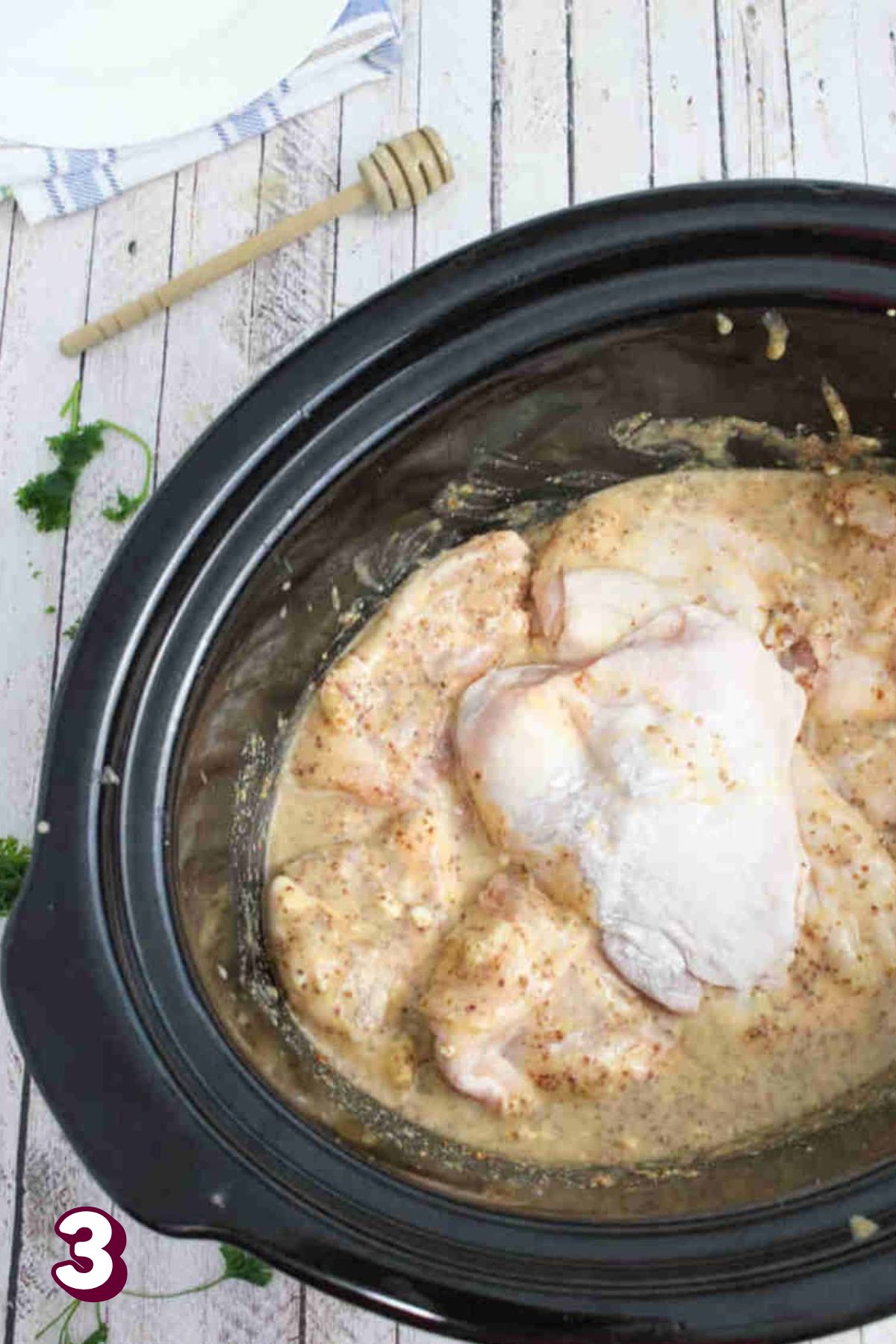 Raw chicken thigh placed on top of others in a honey mustard sauce with more thighs in a black slow cooker.