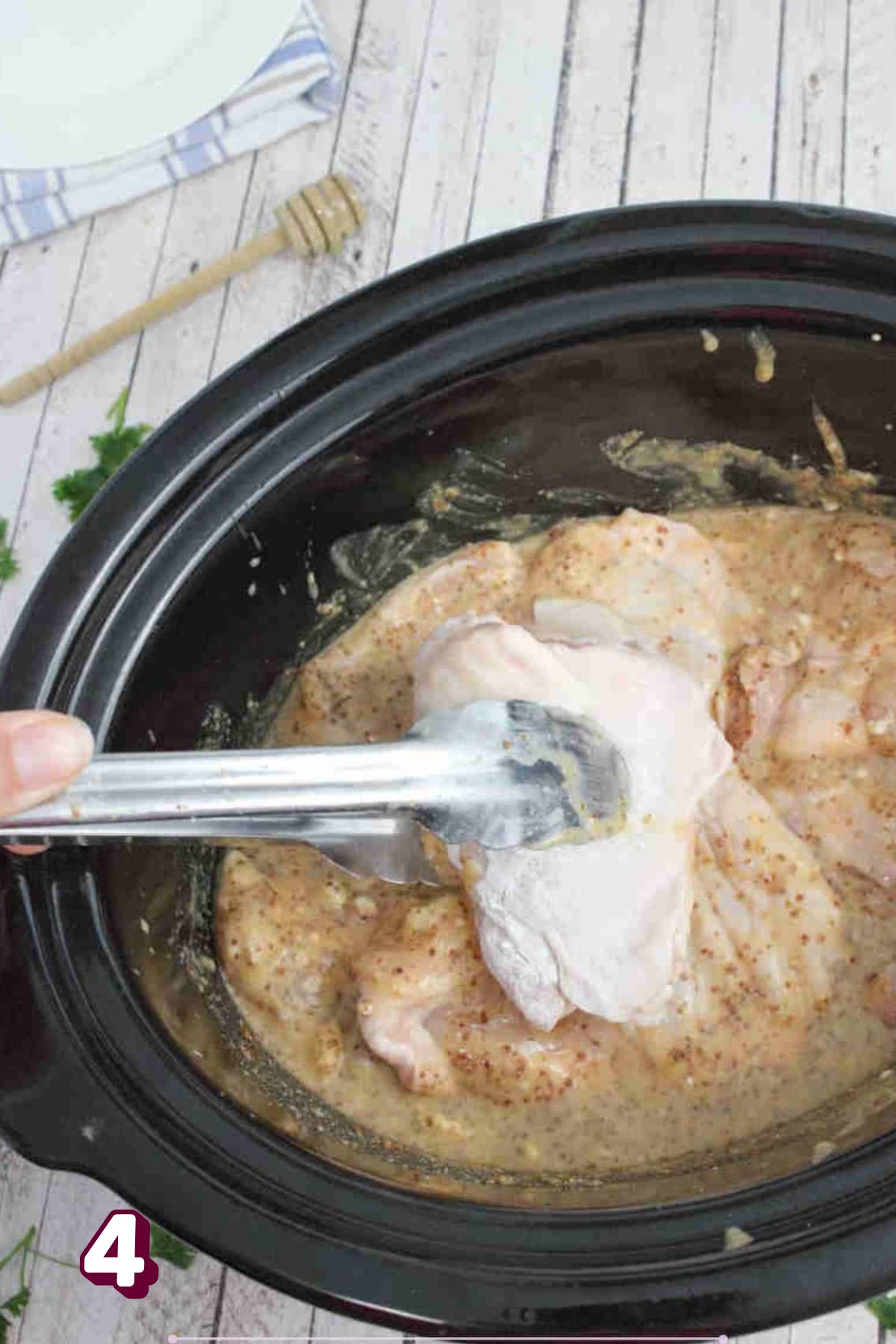 Metal tongs placing a raw chicken thigh into a black slow cooker with honey mustard sauce and other chicken thighs in it.