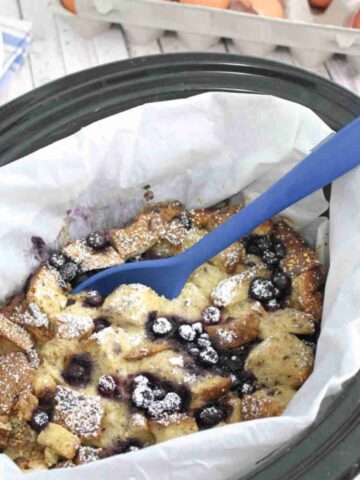 Slow cooker lined with parchment paper and filled with high protein French toast casserole with blueberries with a blue spoon in the bowl.