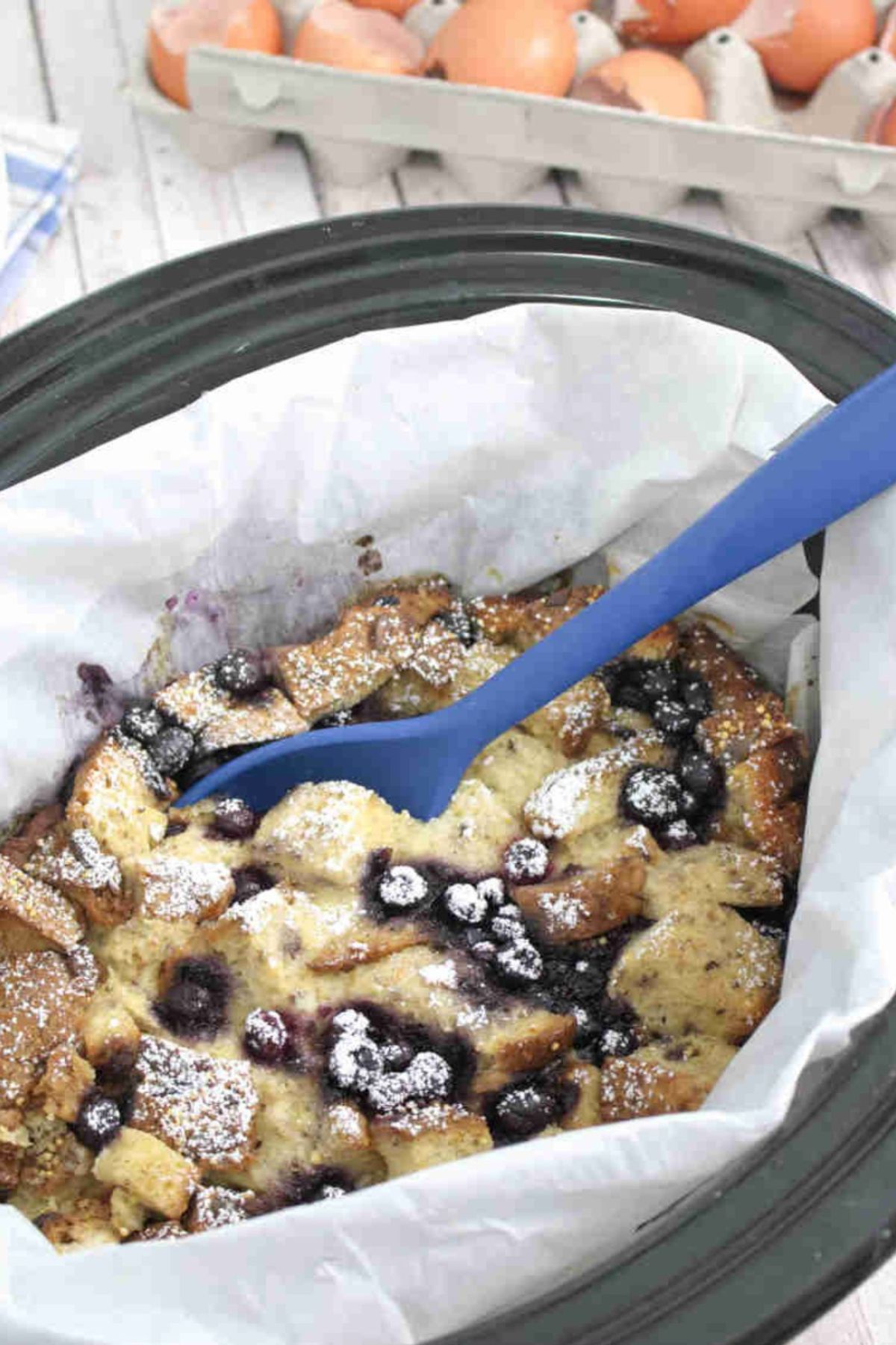 Slow cooker lined with parchment paper and filled with high protein French toast casserole with blueberries with a blue spoon in the bowl.