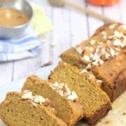 Pumpkin protein bread in slices on a wooden cutting board with almonds, linens, a pumpkin and honey stick around it.